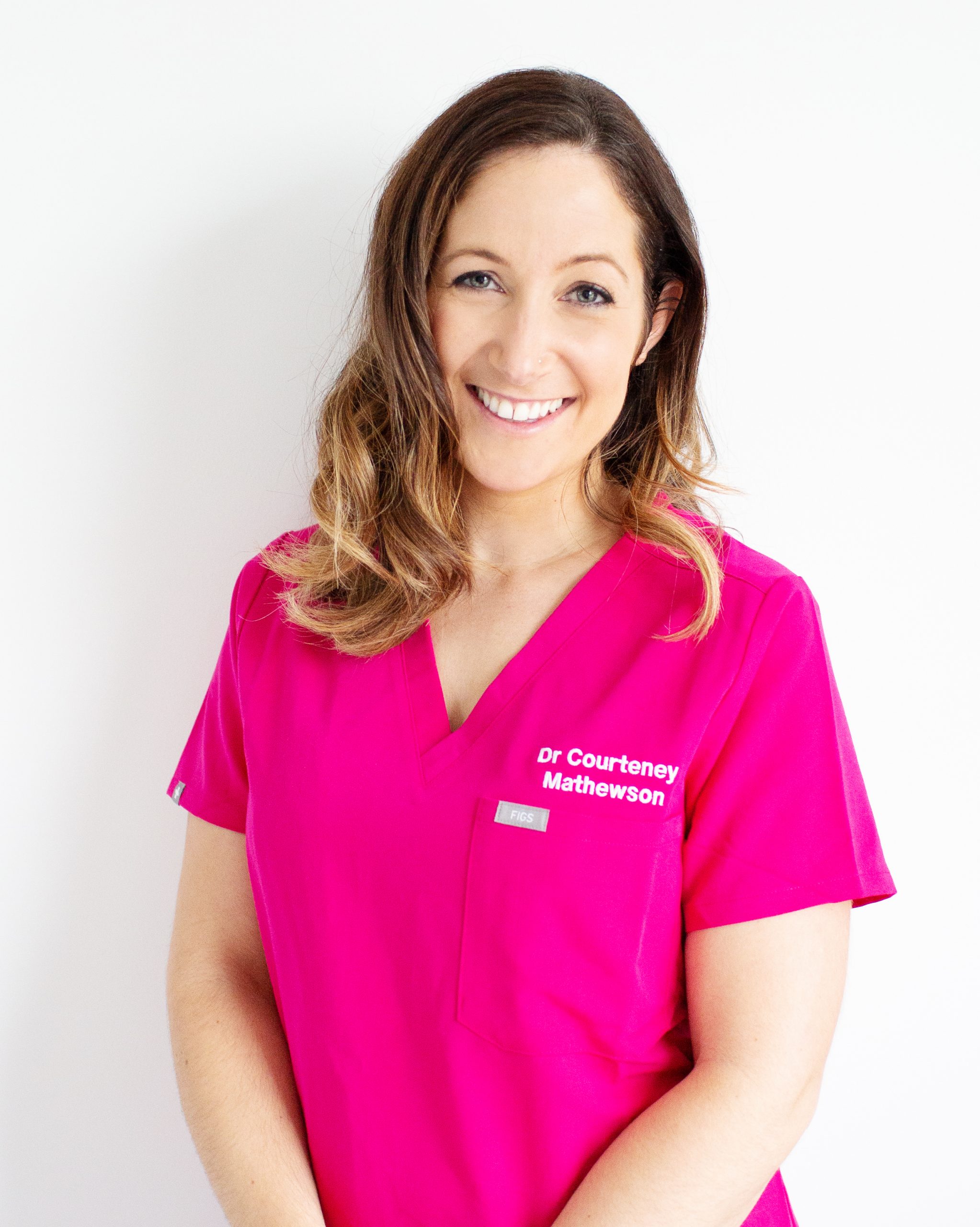 Dr Courteney Mathewson. Face on, pink scrubs, long brown hair curled. Smiling, friendly, empathetic, medical, understanding. Safe and professional.